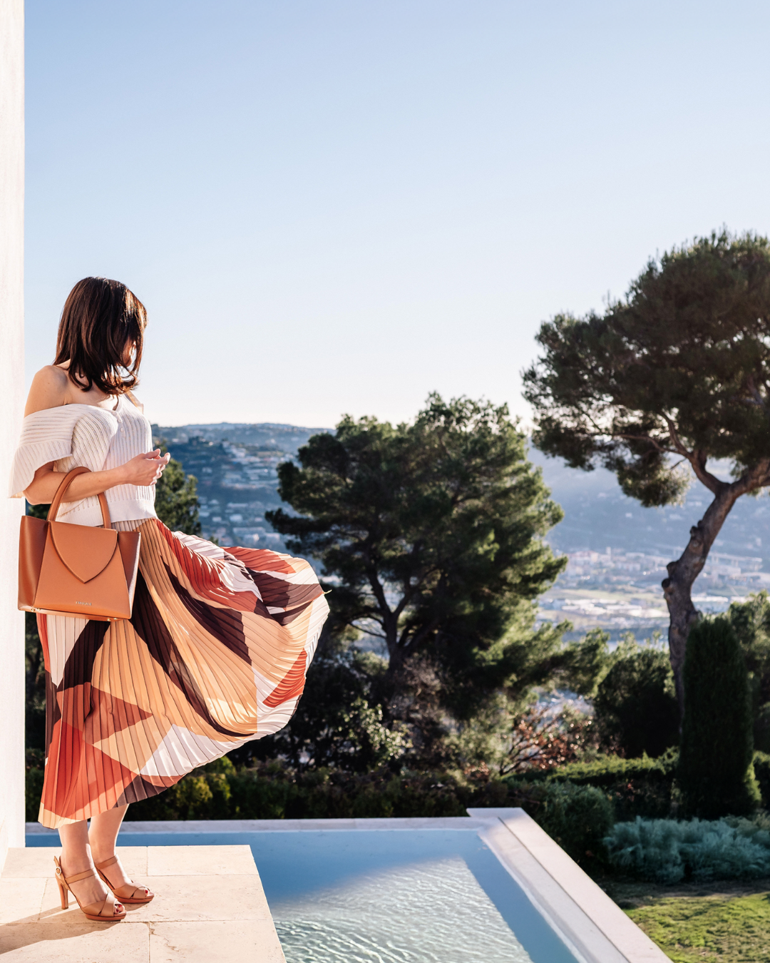 Femme élégante portant le sac Désinvolte couleur miel de FRIVOLE Riviera, près d’une piscine avec vue sur la Méditerranée, incarne le luxe éthique et solaire.