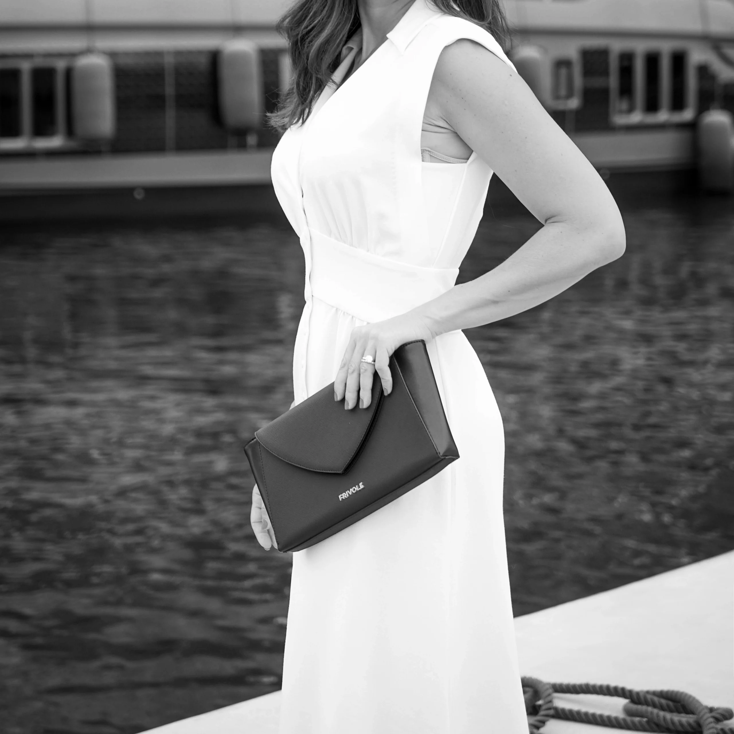 Pochette noire Frivole tenue à la main par une femme en robe blanche, de profil au bord de l’eau.